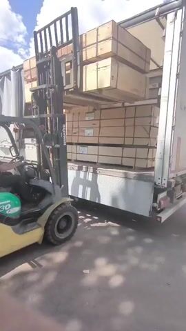 A yellow forklift is loading wooden crates into the back of a white semi-trailer truck. The truck is parked on a paved surface under a partly cloudy blue sky. The forklift is operated by a person wearing a dark shirt and a hat. The forklift's forks are elevated, carrying a stack of wooden crates. The back of the truck is open, revealing rows of stacked wooden crates inside. There are wooden planks on the ground near the back of the truck.