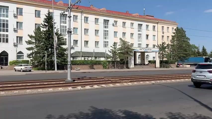 The video captures a wide shot of a large, multi-story building with a red roof. The building is light-colored, possibly cream or beige, with many windows. In front of the building is a street with tram tracks running horizontally across the frame. On the left side of the street, a white car is parked. On the right side of the street, a blue semi-truck is visible, along with other vehicles. Several evergreen trees are planted in front of the building. A utility pole with wires is in the foreground, obscuring a portion of the building and the street. The sky is clear and blue, indicating a sunny day. The overall scene appears to be urban or suburban.