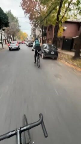 A first-person perspective video follows a person riding a bicycle down a quiet street on an overcast day. The road is paved and surrounded by buildings and trees with autumn foliage. Several parked cars line the street. The cameraperson follows another cyclist who is wearing a light green jacket, black pants with white stripes, and a dark-colored backpack. The cyclist in front also wears a face mask and a baseball cap under a hood. The cyclist ahead glances back towards the cameraperson at one point.