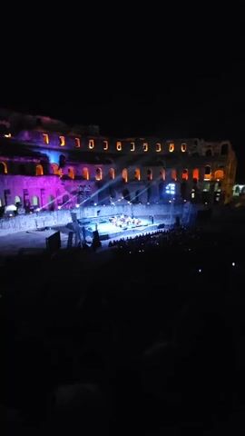 This video shows a nighttime outdoor concert or performance taking place inside an ancient amphitheater. A stage is set up in the center of the arena, illuminated by blue and white spotlights. On the stage, a group of musicians, possibly an orchestra, are performing. They are arranged around a piano and several other instruments, including what appear to be string instruments. The audience is seated in the tiered seating of the amphitheater, with most of them visible in silhouette due to the darkness. The ancient stone structure of the amphitheater is illuminated by purple and orange lights along its upper levels, creating a dramatic backdrop for the performance. The overall atmosphere is that of a lively and visually engaging event.