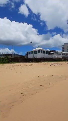 The scene is a beach on a partly cloudy day.  The sand is a golden tan color, and there are a few bits of debris scattered on it.  A red flag on a pole is planted on the sand near the edge of the beach. Beyond the beach, there is some vegetation and a barrier, with buildings and power lines visible further in the distance. The sky is mostly blue with white and grey clouds, creating a mix of bright and shaded areas. The lighting on the beach itself is bright.