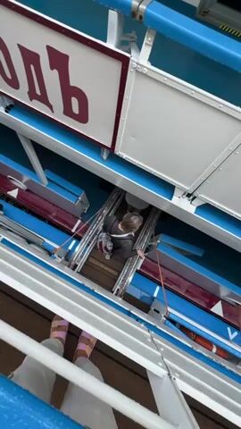 This media file shows a person descending a wooden staircase on what appears to be a boat or ship. The person, wearing a light-colored jacket and dark pants, is moving downwards towards a lower deck. The staircase has metal railings and orange safety lines.

The surrounding environment suggests an outdoor setting, possibly a waterfront or harbor. The side of the vessel is painted blue with red and white accents. A portion of a sign with Cyrillic lettering is visible at the top of the frame. The text, though partially obscured, appears to be "ОДЬ". The weather seems overcast, with visible water droplets on the blue railing in the foreground, indicating it might be raining or recently rained. The overall mood is practical, focusing on the act of movement and the functional aspects of the vessel.

In the foreground, a pair of feet wearing pink and white sandals are visible, suggesting another person is standing on the deck above, looking down at the person descending the stairs. The scene likely takes place in Valaam, Russia, given the context. The time of day is not definitively ascertainable but appears to be daytime due to the ambient light.
