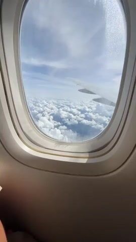 The video shows a woman sitting in an airplane seat by the window. The window offers a view of a sky filled with white clouds. The woman, with her hair tied back, is wearing a light-colored t-shirt and headphones. She is looking out the window with a thoughtful expression. The airplane's interior, including the window frame and a portion of the seat, is visible in the foreground.