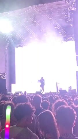 A concert is taking place with a performer on stage in front of a large LED screen. The screen displays an image of the performer, duplicated and with a purple hue. The performer is wearing dark clothing and holding a microphone. There are lights and smoke effects on stage. A crowd of people is visible in the foreground, some holding glowing sticks. The concert appears to be in a large venue, possibly outdoors, with scaffolding and lights visible above the stage.