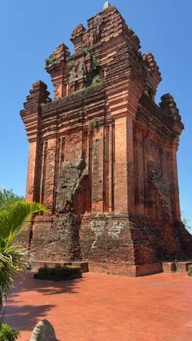 The video shows a close-up, low-angle view of a brick tower under a clear blue sky. A palm tree in the foreground partially obstructs the view of the tower, which is made of red brick and appears ancient and weathered. The tower has multiple tiers and intricate architectural details. The ground in front of the tower is paved with red tiles. A person in a white shirt walks into the frame from the right and is partially visible. Lush green trees and foliage surround the tower. The video pans slightly to the left, revealing more of the surrounding greenery and the blue sky.