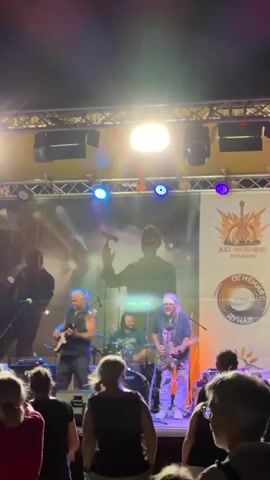 A band is performing on an outdoor stage at night. The band consists of a male bassist on the left, a male drummer in the center, and a male guitarist on the right. There is also a female violinist playing on the far left of the stage, partially obscured by the bassist. The band is playing in front of a large banner that features a silhouette of a person holding a microphone and other graphics. The stage is illuminated by spotlights and colored lights. A crowd of people is visible in the foreground, facing the stage. Some members of the crowd are clapping.