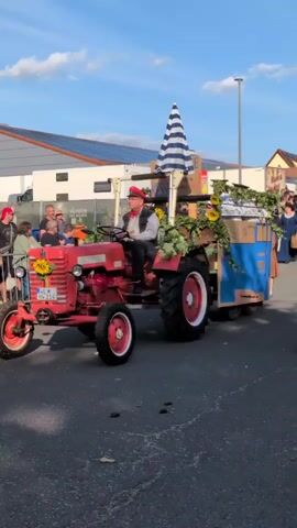 A red McCormick tractor with a man wearing a red cap and vest driving it is moving along a street. The tractor is decorated with sunflowers and greenery. It is pulling a blue and brown trailer also adorned with sunflowers and greenery. A large blue and white striped umbrella is attached to the back of the tractor. Several people are watching the tractor pass by, some are standing on the side of the street and others are on the tractor itself. In the background, there is a yellow building with the words "Buttstädter Vollkornbäckerei Celestina" and some advertisements. The sky is blue with some clouds.