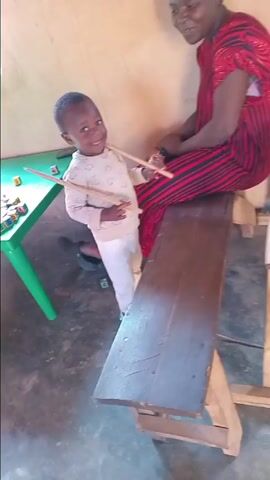 In Barakin Columbai, Nigeria, a young child and an adult woman are engaged in a joyful musical activity indoors. The child, wearing a light-colored long-sleeved top with cloud patterns and light pants, stands enthusiastically in front of a wooden bench. He holds two wooden sticks and uses them to drum on the bench. The woman, dressed in a red and black striped dress, sits on the bench, also holding a wooden stick and a plastic water bottle. She is smiling widely and singing along, using her stick to join the child's drumming. Both individuals are looking at each other, sharing a happy and playful moment. On a green plastic table to the left, several colorful alphabet blocks are visible, displaying letters such as 'E', 'A', 'S', 'M', and 'L'. The room has light-colored walls and a concrete floor. A black slipper is visible on the floor near the child.