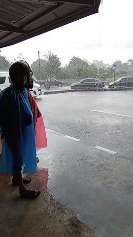 The video shows a rainy scene from under an overhang. Two people wearing ponchos, one blue and one red, stand in the foreground looking out at a street. Rain is visible falling heavily. Cars, including a white van and several sedans, are driving on the wet road. Trees and buildings are visible in the background. The overall atmosphere is wet and overcast.