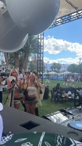 The video shows a lively outdoor event, possibly a festival or party, on a sunny day with palm trees and mountains in the background. Several young women in bohemian-style outfits dance near a DJ booth, holding drinks and fans. A crowd of people mill around, some with water guns and accessories. The camera pans up to reveal a large speaker system and a shaded area with seating. People are talking, laughing, and using their phones, creating a vibrant social atmosphere.