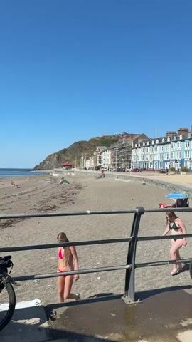A wide shot of a crowded beach on a sunny day with a clear blue sky. In the foreground, a bicycle is parked next to a metal railing. Two young women are visible near the railing, one in a pink bikini and the other in a green bikini. Beyond the railing, the beach is populated with people sunbathing and playing. In the background, there are buildings lining the coast and a hill with some structures on it. The ocean is calm with small waves.