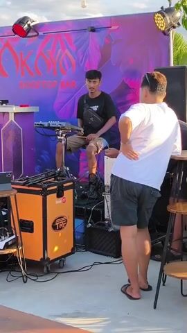 The video shows a rooftop bar with a DJ setup. A man is sitting on a stool playing a percussion instrument, while another man stands beside him. The bar has a backdrop with a floral design and the words "OKAYO ROOFTOP BAR" written on it. There are speakers, lights, and a DJ console visible. In the background, there is a view of mountains and a residential area.