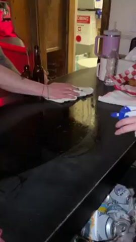 A man with tattooed arms is wiping down a black countertop with a white rag. He is wearing a black baseball cap and a black shirt with a logo on it. There are two bottles of alcohol next to him on the counter. Another person sprays the countertop with a blue and white spray bottle. A water bottle, a mug with a purple handle, and a red and white checkered basket of food are also on the counter. There are cans and other items below the counter.  In the background is a doorway leading to a room with a white box that says "OMING" and has a red cross on it. There is also some writing on a white board. There is a red light above the counter and the bar appears to be busy.