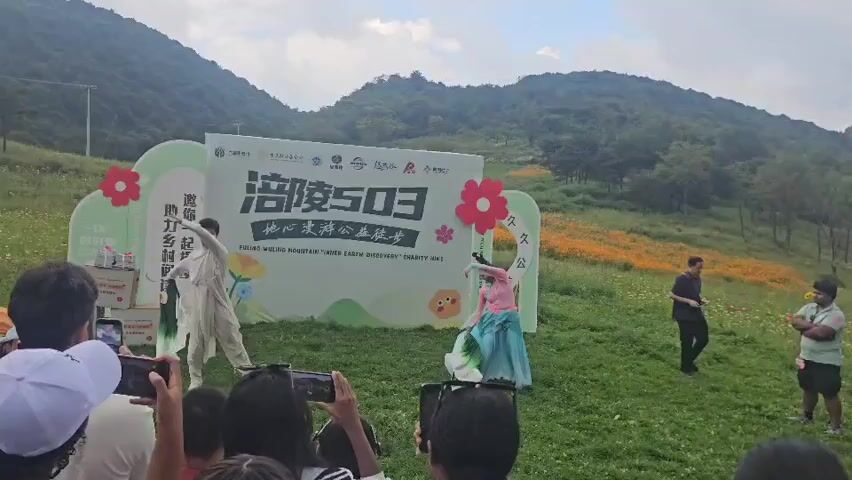 A man in a light-colored costume and a woman in a pink and blue outfit are performing a dance in front of a banner that reads "Hike For Love" and has Chinese characters. The banner is placed in a grassy outdoor area with hills in the background. Several people are watching the performance and recording it with their phones. The man is on the left side of the stage, and the woman is on the right. They are both making graceful movements. There are also some boxes of products on the left side of the stage, and a small table with some items on the right. The sky is cloudy, and there are some trees and yellow flowers visible in the distance.