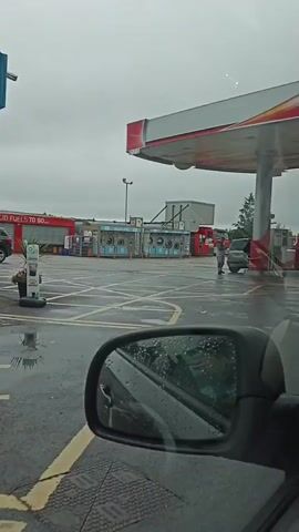 The video shows a view from inside a vehicle looking out at a gas station and convenience store. A black SUV is parked in front of the store, and a person is walking away from it towards a building. The ground is wet, suggesting it is raining or has recently rained. The sky is overcast and gray. There are signs indicating "SOLID FUELS TO GO" and "MACE."