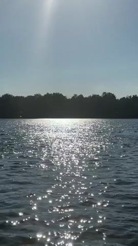 A vertical video captures a serene lake on a sunny day. The sun, high in the sky, casts a bright glare and creates a sparkling reflection on the water's surface, forming a shimmering path that leads towards a silhouetted tree line in the distance. The sky is clear and blue, with the sun's rays creating a lens flare effect in the upper left portion of the frame. The water is calm, with gentle ripples disturbing the reflection. The overall impression is one of peace and natural beauty.