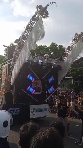 Here is a description of the video:
The video shows a large truck with a massive sound system, adorned with lights, decorative wings, and a portrait of a man. The truck is slowly moving down a street, followed and surrounded by a crowd of people, many of whom are dancing and moving to the music. The people consist of men, women, and children. Some people in the crowd are wearing masks, and some people are dressed casually. The environment includes a suburban street with houses and trees visible in the background. The sky is cloudy.