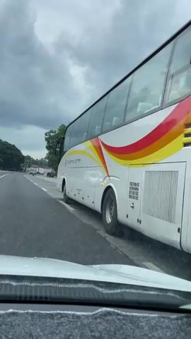 A white, black, red, and yellow Mindanao Star bus is parked on the right side of a gray asphalt road. The bus is angled slightly away from the camera and has its hazard lights flashing. It has dark tinted windows. The bus has the name Mindanao Star printed in large font on its back along with phone numbers. It has a plate number starting with G. A white car is driving on the left lane, and the video is taken from inside this car. The dashboard and top of the hood of the car are visible. The sky is gray and overcast. Trees are on the side of the road. Other cars are in the distance.