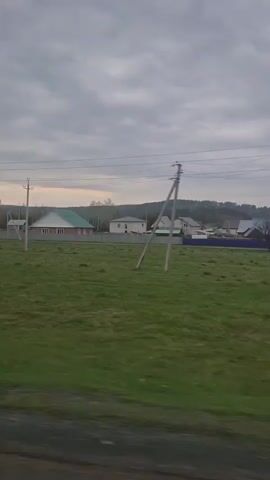 The video shows a view of a rural landscape from a moving vehicle. The sky is overcast with grey clouds. In the foreground, there is a field of green grass with scattered clumps of brown vegetation. Utility poles and power lines run across the scene. In the middle ground, there are several houses, each with a different architectural style and color scheme. Some of the houses have fences around them. Trees are scattered throughout the landscape, and there is a low mountain range visible in the background. There is a small metal utility box in the foreground. The vehicle appears to be traveling along a paved road.