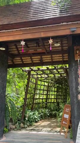 The video begins with a view of an entrance framed by a wooden overhang and decorated with star-shaped ornaments. A path, paved with bricks, leads into a lush green environment covered by a wooden trellis intertwined with plants. To the right, a sign with Thai writing and an arrow pointing right indicates that the establishment is open. The camera then moves forward, following the path under the plant-covered trellis, revealing a serene, green setting with various plants lining the walkway. The path leads to an open, grassy area visible in the distance. The overall impression is one of a peaceful, nature-filled entrance to a garden or outdoor space.