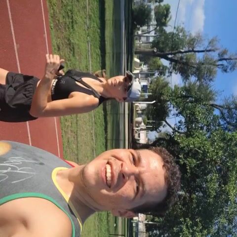 A man and a woman are running on a track. The man is holding a phone and appears to be recording a selfie video while running. He is wearing a grey tank top with a colorful graphic, while the woman is wearing a black sports bra and black shorts. She is holding a phone as well. The track is red with white lines. There is a grassy area next to the track, with trees and a few buildings visible in the background. The sky is blue and clear. The man is smiling, and both appear to be enjoying their run.