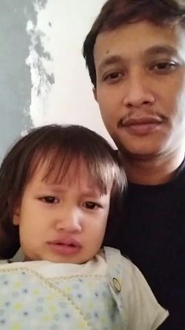 A man and a child are in close proximity, likely taking a selfie. The child, positioned in the foreground, has a distressed expression with tear-filled eyes and a downturned mouth. They are wearing a white shirt with a blue and yellow polka dot pattern. The child's right hand is raised to their face, with their index finger near their nose. The man, in the background, is looking towards the camera with a neutral expression and a mustache. He is wearing a black shirt. The environment appears to be indoors, with a textured, pale wall visible behind them. The lighting suggests an indoor setting. The footage is low quality, with some blur and grain.