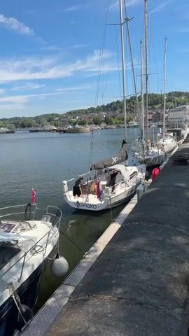 The video shows a view from a dock looking out at several sailboats moored in a harbor. The water is calm and reflects the blue sky with some white clouds. In the foreground, a white sailboat with a black sail is tied to the dock. Two people are visible on the deck of this sailboat, one of whom appears to be sitting. Another sailboat, white with its sail furled, is tied to the first one. Further in the background, more sailboats and other vessels are moored. The dock itself is made of stone or concrete, with a large, dark, rounded mooring bollard visible. Beyond the harbor, a town with buildings and trees lines a gentle hill under the bright sky. The overall scene is peaceful and appears to be a sunny day.