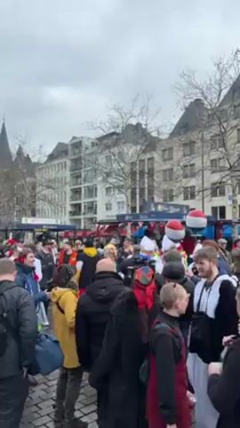 The video shows a crowd of people gathered in a square in front of buildings, with some people wearing costumes. Several food or beverage stands are visible in the background, decorated with bunting. Some individuals are holding red and white balloons. A camera operator with professional equipment is visible, suggesting a possible media presence. The weather appears overcast. People are talking and moving about, and music can be heard in the background.