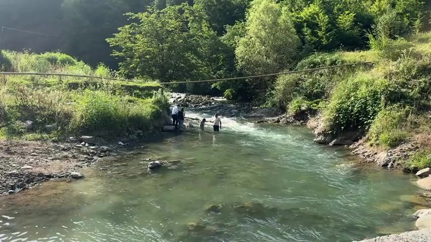 A wide shot shows a group of people standing in a clear, flowing river. The water is shallow enough for them to stand in, with some individuals appearing to be waist-deep. The river is surrounded by lush green vegetation, with trees covering the hillsides in the background. A makeshift bridge made of wire and wood spans the river in the middle ground, with a utility pole visible on the left side of the frame. The sun is shining, casting light on the water and the surrounding landscape. The overall scene depicts a serene natural environment with human activity.