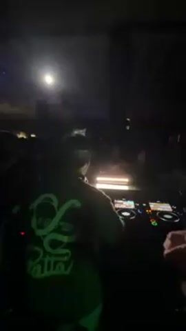 In a dimly lit venue, a DJ in a green Patta hoodie works a DJ mixer while a crowd dances behind them. A spotlight shines on the crowd. Other faces can be seen enjoying the music in the dark background. There are visible support beams on the ceiling. Another person stands to the right side of the DJ mixer.