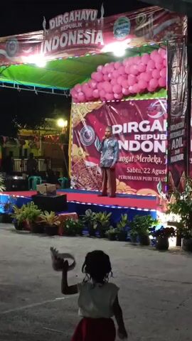 The video captures an outdoor nighttime celebration for Indonesian Independence Day. A stage is set up with a large banner that reads "DIRGAHAYU REPUBLIK INDONESIA" (Long Live the Republic of Indonesia). The stage is decorated with pink balloons and lights. A man is standing on the stage, holding a microphone and speaking. In front of the stage, there are plants in pots and a small child in a red skirt is facing away from the camera, holding something in their hands and looking towards the stage. The background shows some buildings and lights. Another person wearing a white hijab and white top is partially visible in the right foreground, also facing the stage. The overall scene suggests a community event with a festive atmosphere.
