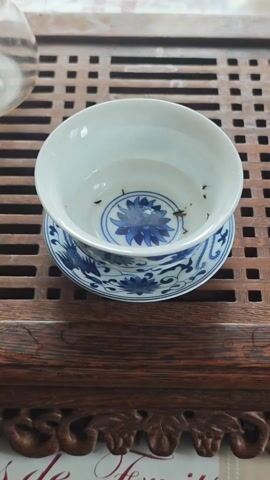A small amount of loose leaf tea is dropped into a white tea bowl that rests on a blue and white patterned saucer, all sitting on a wooden latticed table.