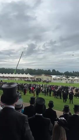 The video shows a horse race from a spectator's perspective. In the foreground, several men are wearing black top hats and formal attire, facing away from the camera and towards the race track. The race track is visible in the mid-ground, with a group of horses and jockeys in mid-stride. A large crowd of spectators, many also in formal wear, are gathered on the grassy area beside the track. In the background, there are large white tents, trees, and a cloudy sky. A tall green crane is visible in the sky over the track.