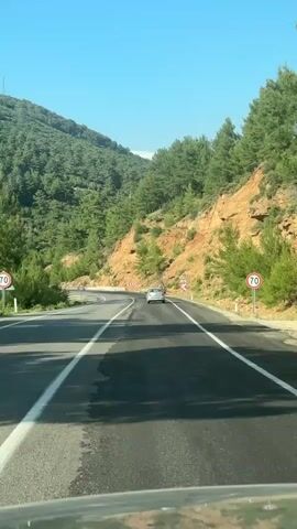A white car is driving on a highway that curves to the left, surrounded by green trees and hills. The car is in the right lane, with a red truck visible in the distance in the left lane. There are speed limit signs and warning signs along the side of the road. The sky is clear and blue.