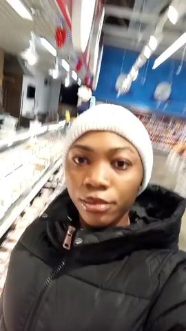 A woman in a white beanie and black puffy coat is filming a selfie video in a grocery store. She appears to be in the meat section, standing in front of a display case filled with various cuts of meat. A worker in a red jacket and hat stands behind the counter, packaging meat. The background includes tiled walls and overhead lighting. The camera moves slightly as the woman films.