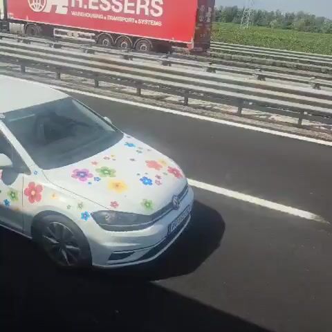 The video shows a white Volkswagen Golf car driving on a highway. The car is decorated with colorful flower stickers on its hood, doors, and rear. The highway appears to be in a rural area, with fields visible in the background. There are other vehicles on the highway, including a white van and a silver car. The video is shot from an elevated perspective, possibly from another vehicle. The weather appears to be sunny.