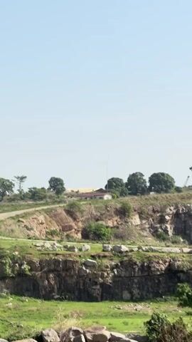 The video shows an overview of a multi-tiered rock quarry on a sunny day. The quarry walls are grey and layered with green vegetation scattered on each level. A small field of green grass is at the base of the quarry. In the distance, a building and some trees can be seen on a hill. The camera pans upwards to show the height of the quarry walls.