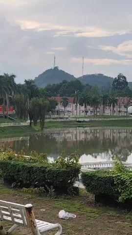 A scenic view of a lake with a reflection of buildings and trees. In the foreground, there is a white bench, some green bushes, and dry grass. The background features a hill with two telecommunication towers and a cloudy sky. The video pans slightly to the right.