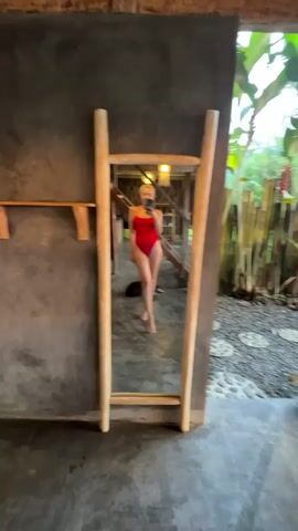 A woman in a red swimsuit stands in front of a full-length mirror, holding up her phone to take a selfie. She is in a room with a dark gray wall, a wooden shelf, and a concrete floor. In the background, through an open doorway, there is a view of lush green vegetation, bamboo fencing, and a cloudy sky. The room appears to be open-air, with a thatched roof. The camera then pans to the right, revealing a wicker chair and a pink yoga mat on the floor. The view of the jungle and sky is more prominent in this shot. The scene is bathed in natural light, suggesting it is daytime, possibly late afternoon given the quality of the light.