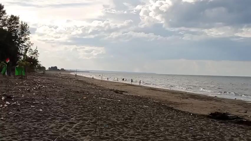 The video shows a beach scene with dark sand, waves crashing on the shore, and a cloudy sky. There are several people walking along the shoreline and a green tent visible on the left side of the frame. Trash is scattered along the beach, including plastic bottles and debris. Some fishing rods are also stuck in the sand.