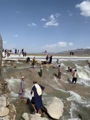 The video captures a scenic outdoor setting featuring a wide, rocky river or stream with numerous people gathered around and within its flowing waters. The scene is bright, under a clear blue sky dotted with scattered white clouds, suggesting a sunny day. In the far background, a range of barren mountains completes the natural landscape.

The river itself is characterized by several tiers of light-colored, uneven rocks over which the water cascades, creating small rapids and white foam. Many individuals, predominantly men and boys dressed in traditional loose-fitting tunics, trousers, and some with head coverings, are actively engaging with the water. Some are standing directly in the shallow, turbulent areas of the river, with water reaching their ankles or calves. Others are carefully navigating or standing on the larger, exposed rocks that dot the riverbed. A young boy in a light tunic and white cap, and a man in a dark blue tunic and white shawl, are visible in the foreground, near the camera, standing in the water.

Along the banks of the river, more people are observed standing and walking, seemingly watching the activity in the water. The overall impression is one of a community or group enjoying the natural environment, possibly for leisure, cooling off, or bathing. The dominant sound throughout the video is the distinct, continuous rush of flowing water.