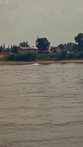 The video shows a wide river with choppy water. In the background, a small village is visible on a hill. The village has several houses and two churches with blue domes. A small boat is seen speeding across the water, leaving a wake. The sky is overcast.