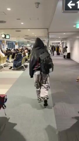 A person wearing a black hoodie, gray cargo pants, and a black Adidas backpack walks down a gray carpeted hallway at an airport. The person is walking away from the camera. To the left, there are rows of chairs filled with people waiting. To the right, other people are walking in the opposite direction. An announcement is being made in Japanese.