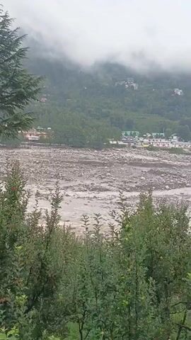 The video shows a wide shot of a landscape with a river in the foreground and mountains in the background. The river appears to be flowing through a valley and is filled with rocks and debris. The mountains are covered in lush green vegetation and are shrouded in mist. There are a few buildings visible on the banks of the river, and some trees are scattered throughout the scene. The overall mood of the video is peaceful and serene.
