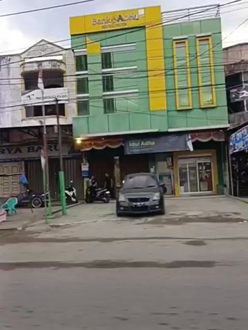 A street scene, possibly in Asia, shows a row of commercial buildings and street vendors. On the right, a yellow and green building displays a sign for "Bank Aceh" and "Idul Adha." In front of it, a dark-colored SUV is parked. To the left, a vendor with a food cart is visible, along with several motorbikes. Further left, a man stands near a stall with red umbrellas, displaying durian fruit. Another vendor sits behind a table laden with goods. A black pickup truck is parked on the street in front of buildings that appear to be shops, one with a sign for "Auto Motor." The sky is overcast.