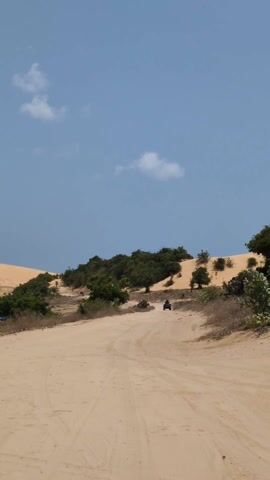 The scene shows a sandy path stretching between dunes and sparse vegetation under a bright blue sky with scattered clouds. An all-terrain vehicle (ATV) is driving towards the camera along the path, kicking up dust.  The ATV has a driver and potentially a passenger. Other ATVs are seen further away on the path. The environment suggests a desert-like landscape.