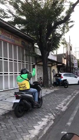 The video shows a street scene with several buildings and vehicles. In the foreground, a motorcycle is partially visible on the left side of the frame. The street is paved with cobblestones. Further down the street, there is a person sitting on the ground next to a tree. The person appears to be wearing a light-colored shirt and dark pants. Behind the person, there is a building with a large rolling metal door that is partially open. Another building to the right has a similar rolling door, which is closed and covered in graffiti. A white car is parked on the right side of the street, and a black motorcycle is parked next to it. The buildings have a commercial appearance, with signs visible above the doors. The overall environment appears to be an urban or semi-urban setting, possibly in a warm climate given the presence of a large tree. The lighting suggests it is daytime, with some shadows indicating the sun is not directly overhead.
