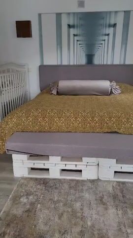 This media file shows a bedroom. The bed is the central object in the scene. It has a grey headboard and a grey mattress, topped with a gold-colored comforter featuring a decorative pattern. A long, cylindrical grey pillow with tied ends rests on the comforter. The bed frame is constructed from white wooden pallets. The room has a modern aesthetic, with a large, panoramic wall mural behind the bed depicting a vanishing-point perspective of a futuristic hallway or tunnel. To the left of the bed, a white crib is visible. To the right, a small white nightstand with decorative detailing and a gold drawer pull is present. A dark, square object is mounted on the wall above the nightstand. The floor is covered with a textured grey rug. The lighting suggests it is daytime, and the overall atmosphere appears calm and tidy. There is no visible text in the media file.
