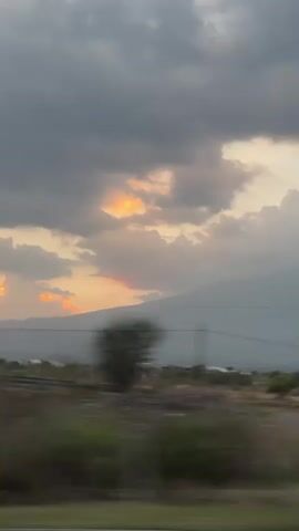 The video shows a view from a moving vehicle, likely a car or bus, looking out towards a rural landscape. The sky is overcast with gray clouds, but there are patches of golden light visible where the sun is breaking through. In the distance, there is a large, hazy mountain. In the foreground, there is sparse vegetation and some scattered buildings. The camera movement is shaky due to the motion of the vehicle. There is no audio.