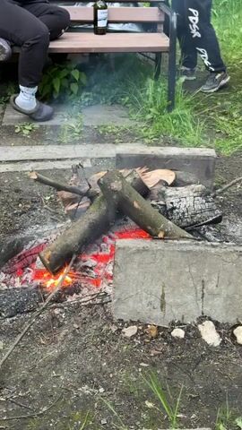 The scene depicts a partially active campfire, with logs partially consumed by flames and embers glowing beneath. The fire is contained within a simple fire pit constructed from four concrete blocks. To the left of the fire pit, a person is seated on a wooden bench. A dark bottle is placed on the bench beside the person. To the right of the fire pit, another person is standing, with only their lower legs and shoes visible. The surrounding ground appears damp, with patches of grass and sparse vegetation scattered around the fire pit. Some smoke emanates from the burning wood.
